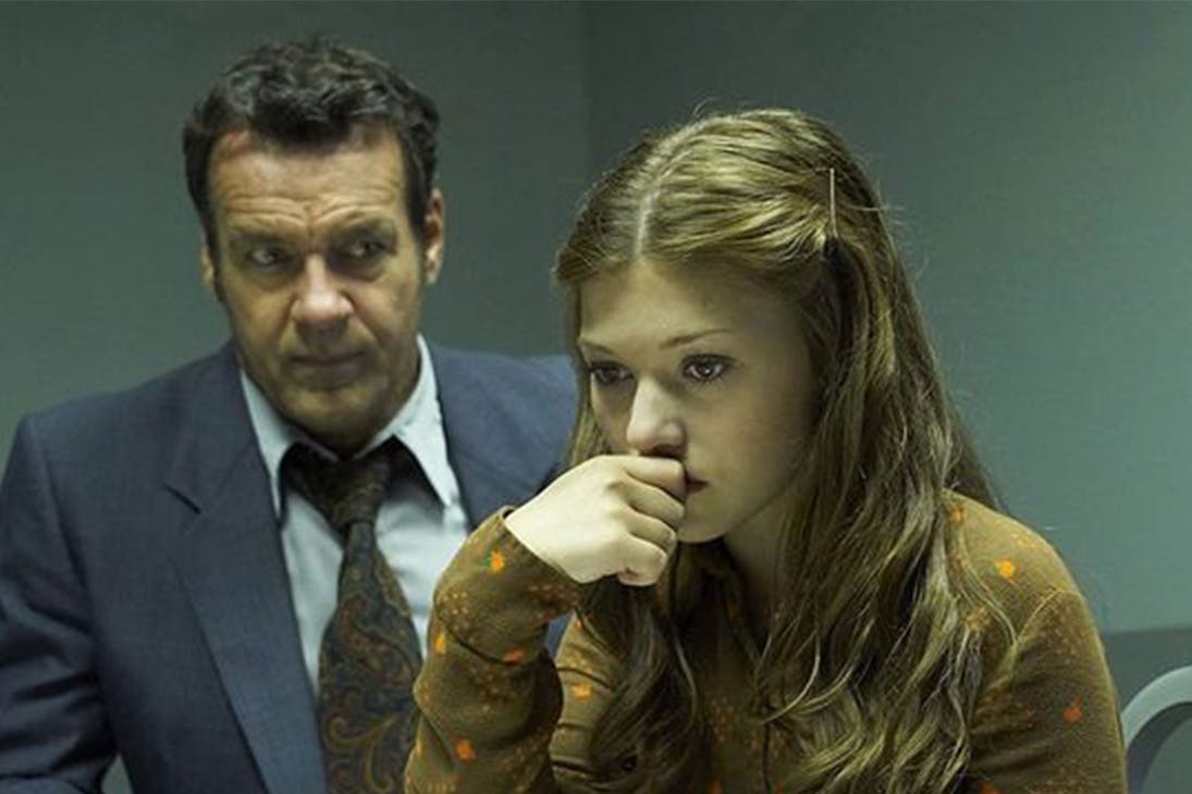 a young woman rests her head on her hand while a man in a suit sits behind her.